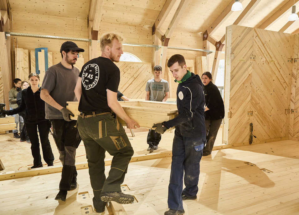 Gruppe von Menschen in einer Holzhütte trägt gemeinsam ein großes Holzbrett