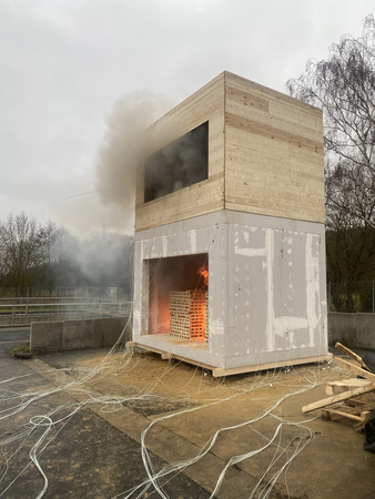 Zweistöckiger Baukörper aus Holz und Gipskartonplatten mit offenem unteren Bereich, in dem ein Feuer brennt und Rauch aufsteigt, umgeben von Kabeln auf dem Boden