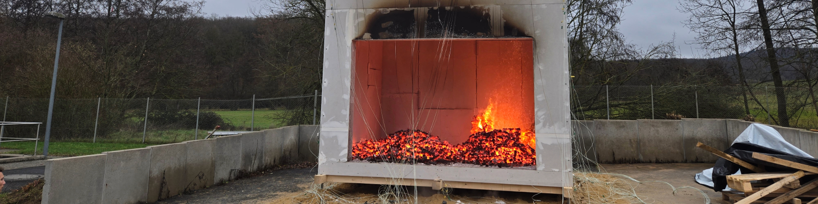 Holztafelkonstruktion mit offenem unteren Bereich, in dem ein Feuer brennt, auf einem Freigelände mit Bäumen im Hintergrund